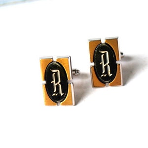 Könnte beinhalten: Ein Paar Manschettenknöpfe aus Gold und Silber mit einem schwarzen ovalen Zentrum, das einen weißen Buchstaben 'R' in einer stilisierten Schrift zeigt.