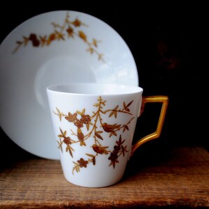 Pode incluir: Uma chávena de chá e um pires brancos com um desenho floral dourado com pássaros. A chávena tem uma pega dourada.