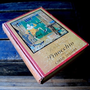 Puede incluir: Libro de tapa dura vintage titulado "Pinocchio" de Carlo Collodi. La portada presenta una colorida ilustración de un personaje con un atuendo amarillo y naranja. El libro tiene una lomo rojo y está sobre una superficie de madera oscura.