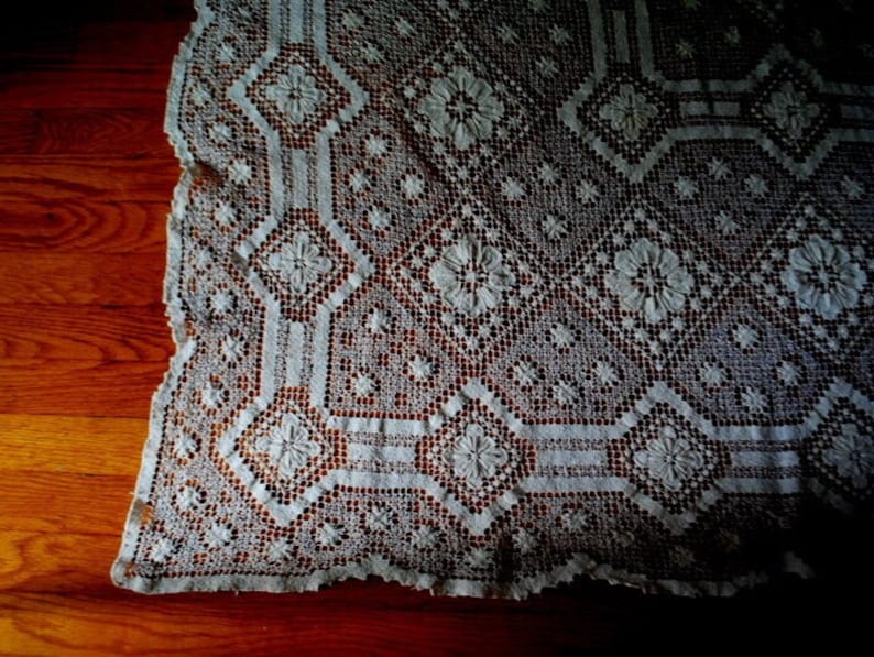 K&ouml;nnte beinhalten: Eine wei&szlig;e H&auml;keltischdecke mit einem zarten Blumenmuster. Die Tischdecke ist aus feiner Spitze und hat einen gewellten Rand.