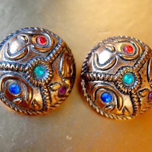 Op de afbeelding: Een paar gouden oorbellen met ingewikkelde ontwerpen en gekleurde edelstenen. De oorbellen zijn rond en hebben een reliëf, getextureerd oppervlak. De edelstenen zijn rood, blauw en groen.
