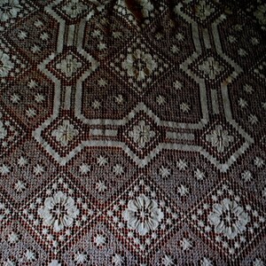 K&ouml;nnte beinhalten: Nahaufnahme einer wei&szlig;en H&auml;keltischdecke mit einem zarten Blumenmuster. Die Tischdecke hat einen dunkelbraunen Hintergrund, der einen Kontrast zwischen den hellen und dunklen Farben erzeugt.