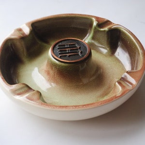 May include: A green and brown ceramic ashtray with a copper-colored center piece. The center piece has a raised design of three vertical lines.