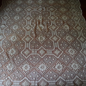 K&ouml;nnte beinhalten: Wei&szlig;e H&auml;keltischdecke mit geometrischem Muster. Die Tischdecke hat ein zartes, filigranes Design mit einem sich wiederholenden Muster aus Rauten und Blumen.