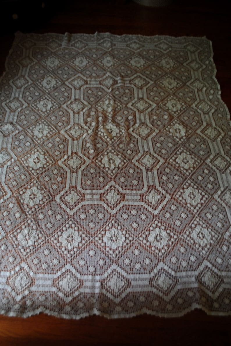 K&ouml;nnte beinhalten: Wei&szlig;e H&auml;keltischdecke mit geometrischem Muster. Die Tischdecke hat ein zartes, filigranes Design mit einem sich wiederholenden Muster aus Rauten und Blumen.