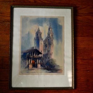May include: A framed watercolor painting depicting a historic stone church with two tall towers and a covered entrance. The artwork features soft blue and tan tones, set within a simple dark frame and a wide white mat board border.