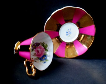 Könnte beinhalten: Ein rosa-goldenes Tee-Set mit Blumenmotiven. Die Untertasse hat einen breiten Rand mit abwechselnden rosa und goldenen Streifen. Die Teetasse hat ein Blumenmuster mit rosa, weißen und gelben Blumen.