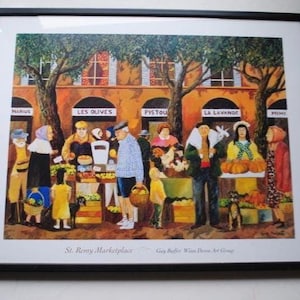 May include: A framed print of a bustling marketplace scene. The artwork depicts various vendors and customers in front of shops with signs. The text "St. Remy Marketplace" is visible at the bottom. The painting has a warm color palette.