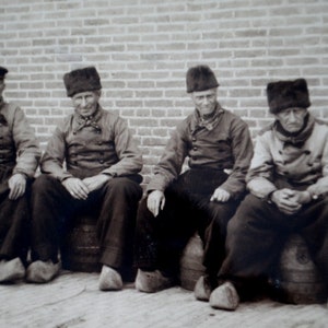 May include: Four men in vintage clothing and hats pose for a photo. They are wearing dark jackets and pants, and some have hats with brims. The men are standing in front of a brick wall.