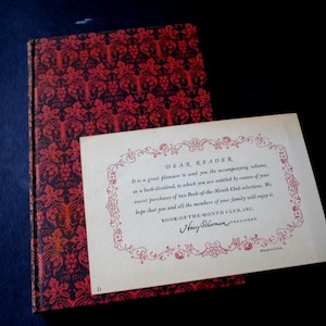 May include: A red and black book with a decorative cover and a white card with the text "Dear Reader, It is a great pleasure to send you the accompanying volume, as a book dividend, to which you are entitled by reason of your recent purchase of two Book-of-the-Month Club selections. We hope that you and all the members of your family will enjoy it." and a signature that reads "Harry Scherman, President, BOOK-OF-THE-MONTH CLUB, INC."
