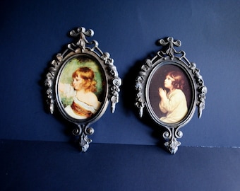 May include: Two ornate silver-toned picture frames with oval-shaped glass panels. Each frame features a detailed floral design and a small, hanging loop at the top. The frames contain vintage-style portraits of children.