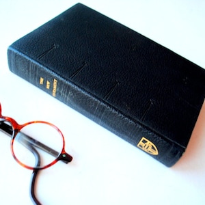May include: A black leather-bound book with gold lettering that reads "NEW TESTAMENT" and a gold emblem on the cover. A pair of tortoiseshell glasses with round lenses are lying on a white surface next to the book.