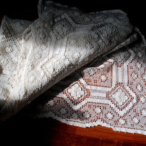K&ouml;nnte beinhalten: Eine wei&szlig;e H&auml;keltischdecke mit einem zarten Blumenmuster. Die Tischdecke ist in der Mitte gefaltet, wodurch die filigranen Details der H&auml;kelarbeit sichtbar werden.