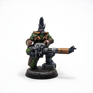 Puede incluir: Figura en miniatura de un personaje con un atuendo verde y marrón, sosteniendo un arma grande y detallada. La figura tiene una cresta y una máscara de respiración. El arma tiene un cañón dorado y está montada sobre una base.