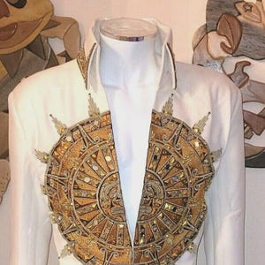 Puede incluir: Mono blanco con adornos dorados y bronce. La chaqueta presenta un gran diseño de sol en el pecho. El cinturón y los pantalones también están adornados con detalles dorados y bronce. El atuendo se exhibe en un maniquí.