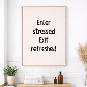 以下が含まれることがあります： 「Enter stressed Exit refreshed」の文字が黒字で書かれた額入りのプリント。白いタオル、鉢植えの植物、茶色のボトルが置かれた木製の棚の上に飾られています。