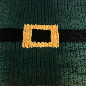 Könnte beinhalten: Nahaufnahme einer handgefertigten grünen, schwarzen und gelben Decke. Die Decke hat ein Design, das einem Gürtel und einer Schnalle ähnelt. Die Textur der Decke erscheint weich und plüschig, mit einem dicken, gestrickten Aussehen. Das Farbschema erinnert an ein festliches Thema.