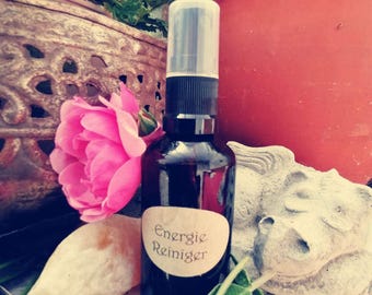 May include: A dark glass spray bottle with a clear cap and a label that reads "Energie Reiniger." The bottle is surrounded by a pink rose, herbs, and a stone sculpture. The background includes a terracotta pot and greenery.