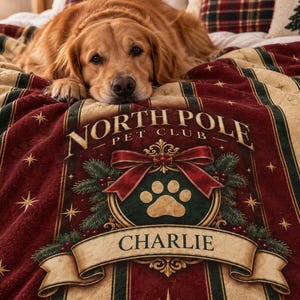May include: A plush blanket featuring a deep red and cream striped pattern with gold star accents. The center displays a crest with a paw print, a red bow, pine branches, and the text "NORTH POLE PET CLUB" above the name "CHARLIE" on a banner.