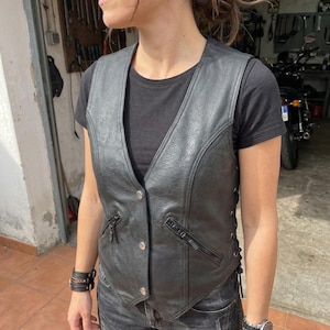 Könnte beinhalten: Schwarze Lederweste mit V-Ausschnitt, Knopfverschluss und Reißverschlusstaschen. Die Weste hat seitliche Schnürung und wird über einem schwarzen T-Shirt und Jeans getragen. Eine Uhr und ein Armband sind sichtbar.