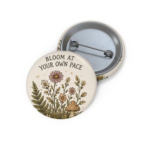 May include: A round pin-back button with a cream-colored background featuring a botanical illustration of wildflowers, a fern, and a mushroom. The text "BLOOM AT YOUR OWN PACE" is printed in black above the floral design. The back shows a metal pin mechanism.