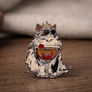 May include: A metal enamel pin featuring a fluffy cat wearing a gold crown and black sunglasses. The cat is holding a cocktail glass filled with an amber liquid, a single red cherry, and ice cubes, rendered in black, white, gold, and red.
