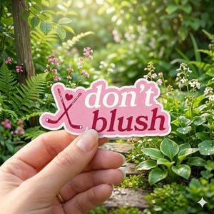 May include: A pink sticker with the words "don't blush" in white and burgundy script. Two crossed hockey sticks and a heart are on the left side. The sticker is held in front of a lush green garden.
