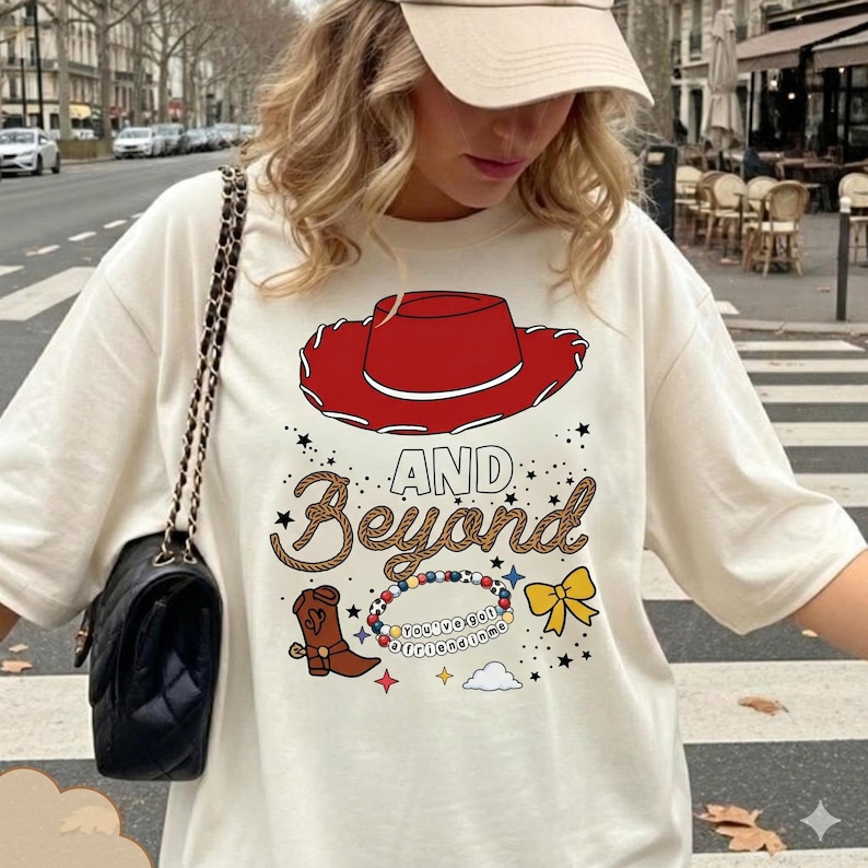 May include: An off-white t-shirt featuring a graphic print of a red cowboy hat, a brown cowboy boot, a yellow bow, and a beaded bracelet with the text 'You've got a friend in me'. The design includes the words 'AND Beyond' in brown rope-style font.