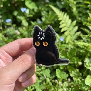 May include: A black cat-shaped acrylic pin featuring a white loading icon on its forehead and two gold-colored circular eye accents. The pin has a clear border and a smooth, glossy finish, depicting a stylized feline silhouette in a sitting pose.
