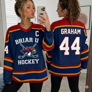 Könnte beinhalten: Marineblaues Hockey-Trikot mit roten und goldenen Akzenten. Das Trikot zeigt den Text "BRIAR U HOCKEY" und ein Logo mit einem Vogel und gekreuzten Hockeyschlägern. Die Nummer "44" und der Name "GRAHAM" sind auf dem Rücken.
