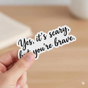 May include: A white sticker with black cursive text that reads "Yes, it's scary, but you're brave." The sticker is held by a hand against a blurred background.