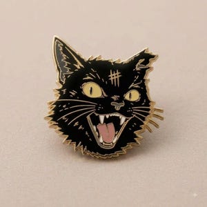 May include: A black enamel pin shaped like a cat head with gold-colored metal outlines. The cat has wide yellow eyes, a pink tongue, sharp white teeth, and a hash mark symbol on its forehead. The design features jagged fur edges.
