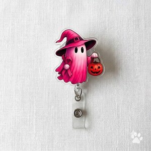 May include: A pink ghost badge reel featuring a character wearing a dark pink witch hat and holding a small orange jack-o'-lantern bucket. The ghost has two black oval eyes and is attached to a clear plastic clip with a retractable cord mechanism.