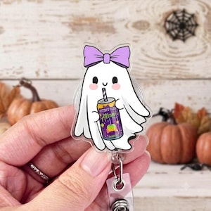 May include: An acrylic badge reel featuring a white ghost character with a light purple bow on its head. The ghost has a smiling face with pink cheeks and is holding a purple energy drink can labeled Witch's Brew Alani with a green apple graphic.