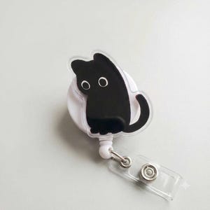 May include: A white retractable badge reel featuring a clear acrylic topper with a black cat illustration. The cat has a solid black body and large white circular eyes. The reel includes a clear plastic strap with a metal snap button attachment.
