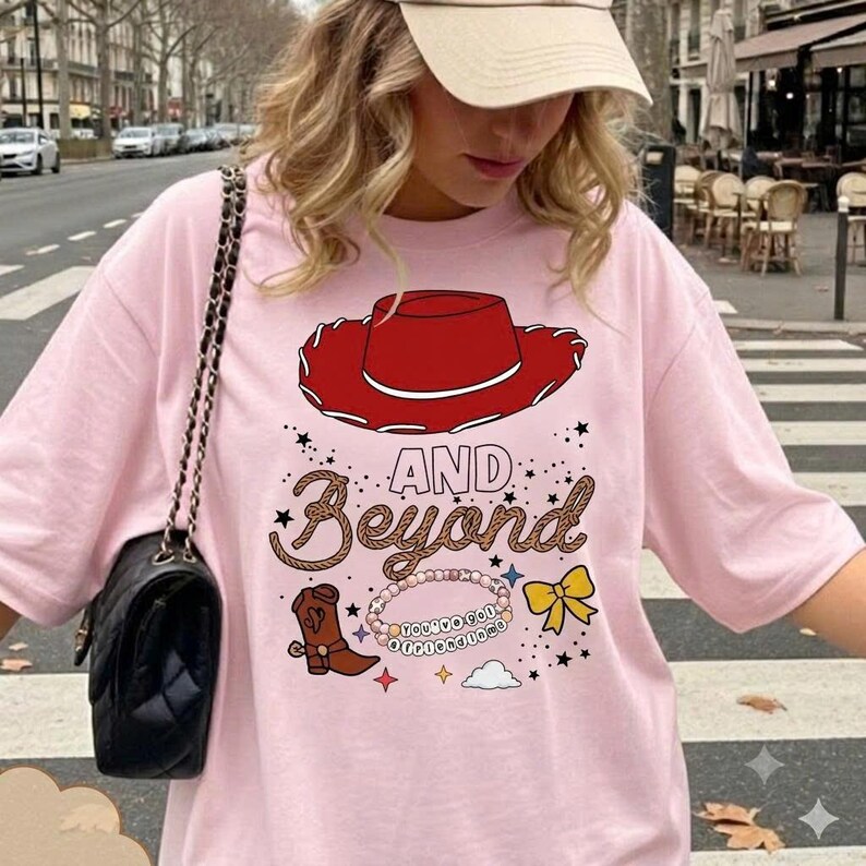 May include: A light pink t-shirt featuring a graphic print of a red cowboy hat, a brown cowboy boot, a yellow bow, and a beaded bracelet with the text "YOU'VE GOT A FRIEND IN ME" above the words "AND BEYOND" in a rope-style font.