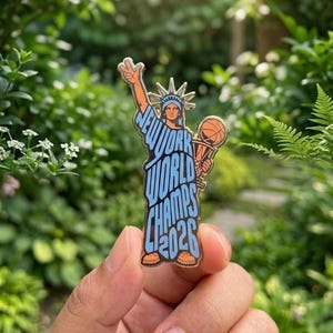 May include: A metal enamel pin shaped like the Statue of Liberty holding a basketball trophy, featuring the text NEW YORK WORLD CHAMPS 2026 in light blue lettering against a dark outline, with orange accents on the shoes and basketball.