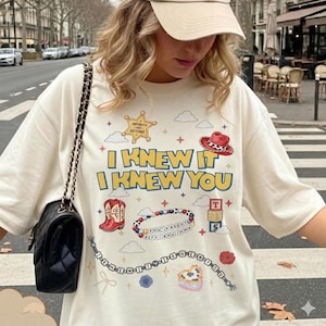 Puede incluir: Una camiseta color crema con un estampado gr&aacute;fico que incluye el texto I KNEW IT I KNEW YOU, una estrella de sheriff, una bota vaquera roja, una pulsera de cuentas, bloques de letras, estrellas y nubes. Se lleva un bolso negro acolchado.