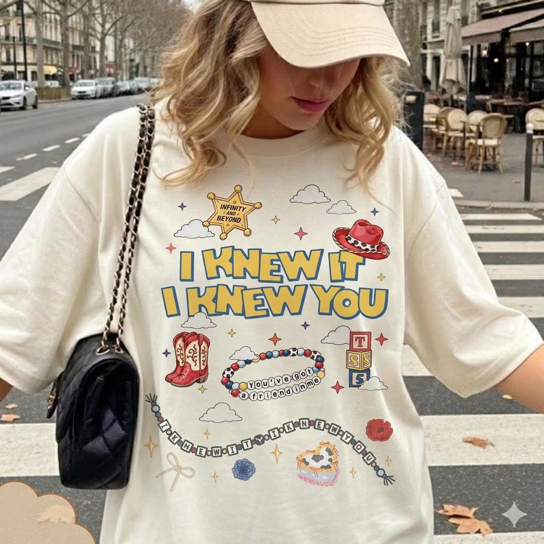 Puede incluir: Una camiseta color crema con un estampado gr&aacute;fico que incluye el texto I KNEW IT I KNEW YOU, una estrella de sheriff, una bota vaquera roja, una pulsera de cuentas, bloques de letras, estrellas y nubes. Se lleva un bolso negro acolchado.
