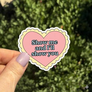 May include: A heart-shaped sticker with a pink center and a scalloped yellow border. The sticker features the text "Show me and I'll show you" in a light blue font. The sticker is held against a green, leafy background.