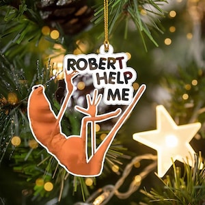 Op de afbeelding: Kerstversiering met een cartoonachtige oranje figuur met uitgestrekte armen. De versiering heeft de tekst "ROBERT HELP ME" in een witte en rode letter. De versiering hangt aan een gouden draad.