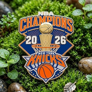 Puede incluir: Un pin de esmalte metálico con el texto NBA CHAMPIONS 2026, un trofeo de baloncesto dorado y un balón. El diseño incluye el logotipo de los New York Knicks en colores azul, naranja y blanco con bordes metálicos dorados y detalles de estrellas.
