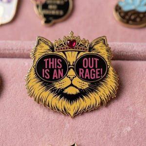 May include: A gold-toned metal enamel pin shaped like a cat face wearing a crown with a red heart gem and black sunglasses. The lenses feature the pink text THIS IS AN OUT RAGE! The cat has yellow fur with black line details and a black nose.