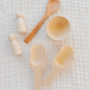 May include: A collection of wooden toys, including two peg dolls, a spoon, a small bowl, and two scoops. The items are made of light-colored wood and are arranged on a white, textured surface. The spoon and bowl have a warm, natural wood tone.