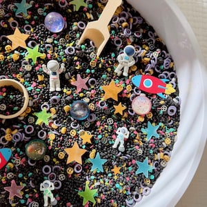 May include: A white circular sensory bin filled with black filler beads, small colorful star shapes, miniature astronaut figurines, a red and blue rocket ship, translucent spheres, and tiny gear-shaped pieces, accompanied by a small wooden scoop.