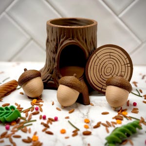 May include: A brown tree stump container with a hollow opening, accompanied by three small acorn-shaped objects with brown caps and tan bases, and a circular flat piece featuring a brown wood grain pattern on a white surface.