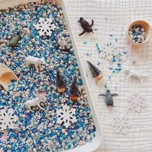 May include: A white rectangular tray filled with blue, white, and pearl-colored sensory materials. Miniature toy animals, including a polar bear, penguin, and moose, are scattered throughout. Decorative snowflakes and small Christmas trees are also present.