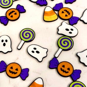 May include: A collection of flat Halloween-themed charms featuring white ghosts with black eyes and mouths, orange jack-o'-lantern candies with purple wrappers, green and purple swirl lollipops, and yellow, orange, and white candy corn pieces.