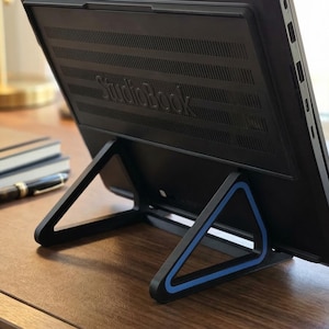 May include: A black laptop computer is propped up on a black stand with blue accents. The stand is triangular and supports the laptop at an angle. The laptop has the word "StudioBook" printed on the back.