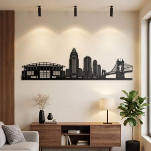 Può includere: Un'opera d'arte da parete in metallo nero che presenta una silhouette dettagliata dello skyline di Cincinnati, inclusi lo stadio, grattacieli prominenti e il ponte sospeso John A. Roebling, montata orizzontalmente su una parete interna.